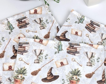 May include: Three white fabric napkins with a whimsical pattern of books, potions, brooms, and witch hats. The pattern is in shades of brown, green, and gold on a white background.