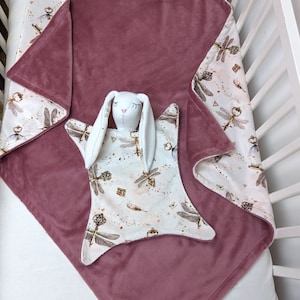 Könnte beinhalten: Eine Babydecke in Staubrosa mit einem weißen und goldenen Libellenmuster an den Rändern. Ein passendes Kuscheltuch-Kaninchen mit langen Ohren und geschlossenen Augen liegt obenauf und bildet ein gemütliches Kinderzimmer-Set.