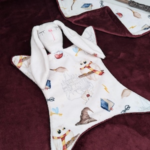 May include: A white bunny security blanket with a white plush bunny head and long ears. The blanket is printed with a white and gray Hogwarts castle, owls, wands, and other Harry Potter themed imagery.