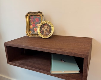 May include: A dark brown floating shelf with a book and two gold picture frames. The picture frames have a floral design and a picture of a dog.