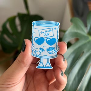 Pode incluir: Um adesivo azul e branco com uma lata de xarope de bordo de desenho animado com óculos de sol em forma de coração. A lata tem braços e pernas e está segurando mirtilos. O texto "Sirop d'érable PUR" está escrito na lata.