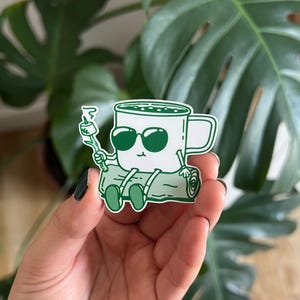 Pode incluir: Um autocolante verde com uma caneca de café de desenho animado a usar óculos de sol e sentada num tronco, segurando um marshmallow num pau. O autocolante tem um contorno branco. Fundo de folhas verdes.