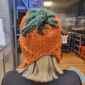 May include: A knitted pumpkin hat with a green top and orange body. The hat is being worn by a person with blonde hair.