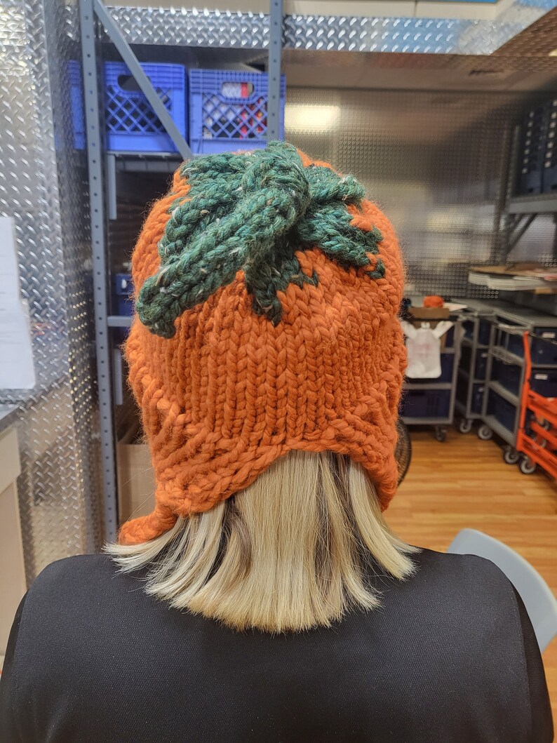 May include: A knitted pumpkin hat with a green top and orange body. The hat is being worn by a person with blonde hair.