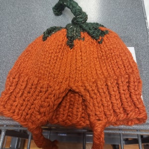 May include: A knitted orange pumpkin hat with a green stem. The hat has two long ties that can be tied under the chin.