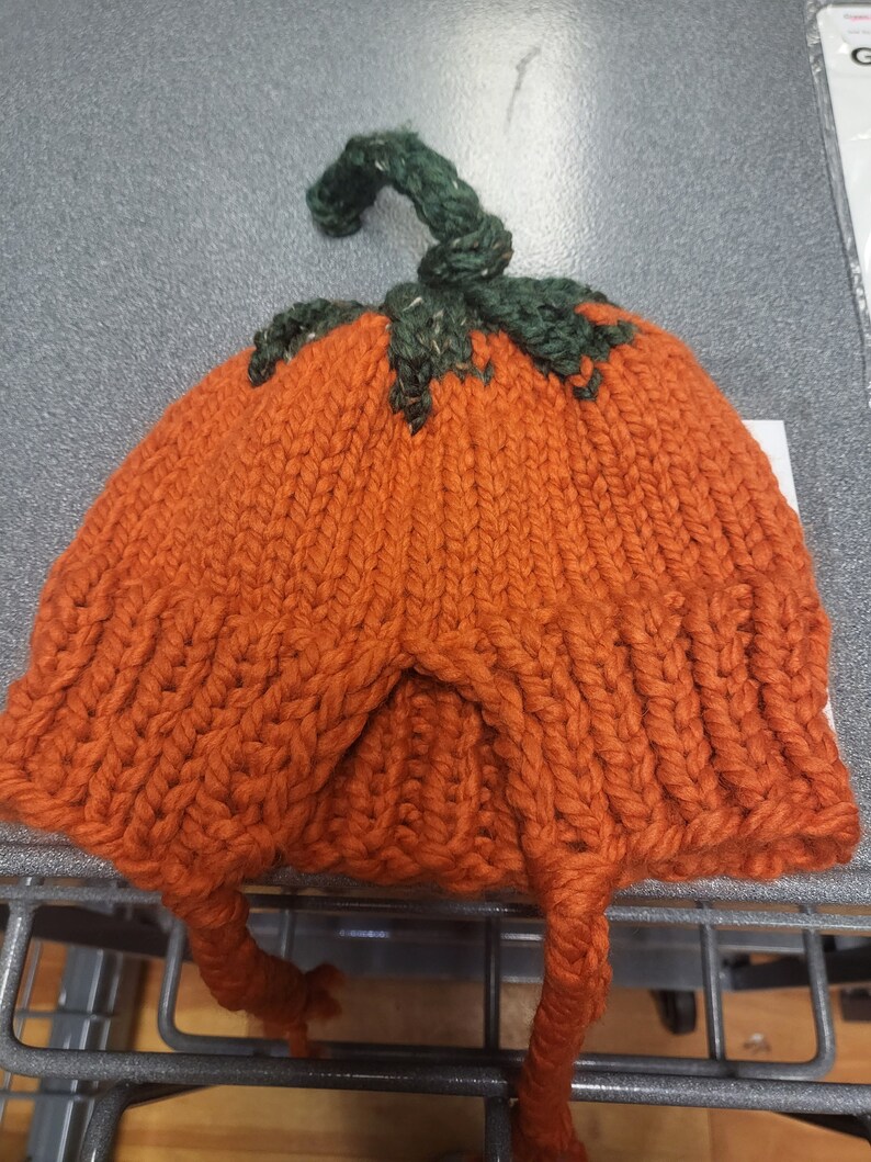May include: A knitted orange pumpkin hat with a green stem. The hat has two long ties that can be tied under the chin.