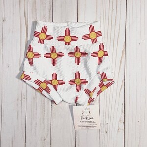 May include: White baby shorts with a repeating pattern of the New Mexico Zia symbol in red and yellow. The shorts have a textured fabric and a high waistband. A small tag with text is visible.