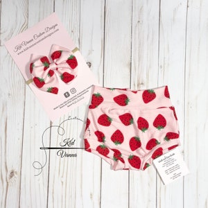 Può includere: Un paio di bloomers per neonati rosa e rossi con stampa a fragole, con un cerchietto abbinato rosa e rosso con un fiocco a forma di fragola. Il testo "Kid Vanna Custom Designs" e "www.kidvannacustomdesigns.com" è su una carta bianca.