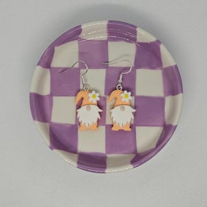 Puede incluir: Un par de pendientes colgantes con un diseño de gnomo. Los gnomos son de color melocotón con barbas y sombreros blancos. Cada gnomo tiene una sola flor blanca en su sombrero. Los pendientes están sobre un plato a cuadros blanco y morado.