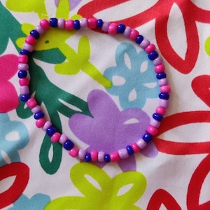 Könnte beinhalten: Ein Armband aus rosa, lila und blauen Perlen. Das Schmuckstück wird vor einem Hintergrund aus Stoff mit einem Blumenmuster mit roten, grünen und lila Blumen präsentiert.