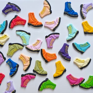 Może przedstawiać: Kolekcja kolorowych, miniaturowych ozdób w kształcie butów. Buty są wykonane z różnych materiałów, w tym filcu, tkaniny i plastiku. Są ułożone w losowym wzorze na białym tle.