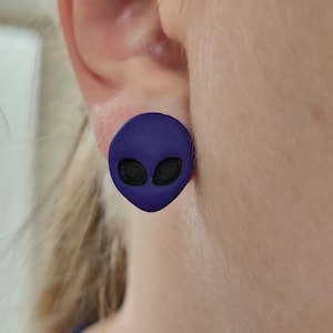 Puede incluir: Pendientes de botón con forma de cara de extraterrestre de color morado. Los pendientes están hechos de un material suave y flexible y presentan una cara de extraterrestre negra con ojos grandes.