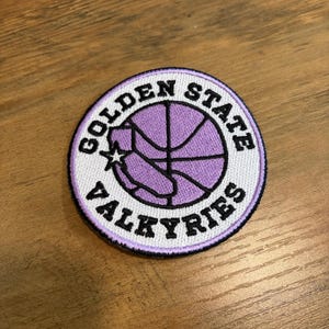 May include: A circular embroidered patch featuring a lavender basketball graphic with a small white star accent. The text GOLDEN STATE VALKYRIES is stitched in black capital letters along the white border, which is finished with a dark purple edge.