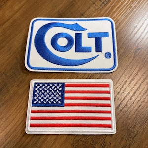 May include: Two embroidered patches. The top patch features the word "COLT" in blue, with a blue logo on a white background. The bottom patch is a rectangular American flag with red and white stripes and a blue field of stars.