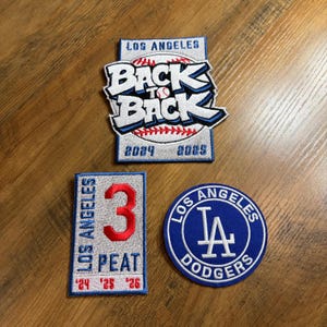 May include: Three embroidered patches featuring the Los Angeles Dodgers logo and baseball-themed designs. One patch reads "Back to Back" with the years 2024 and 2025. Another says "3 Peat" with the years 2024, 2025, and 2026. The third patch is a blue circle with the Dodgers logo.