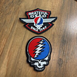 May include: Two embroidered patches. The top patch is a black shield shape with white wings, a red border, and the text "GRATEFUL DEAD SINCE 1965" surrounding a skull icon. The bottom patch is a circular skull design with red and blue sections.