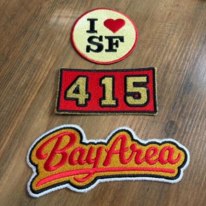 May include: Three embroidered patches. The top is a circular cream patch with a red heart and black text reading I heart SF. The middle is a rectangular red patch with gold numbers 415. The bottom is a gold script patch reading Bay Area in red.