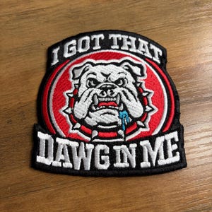 May include: Embroidered patch featuring a bulldog with the text "I GOT THAT DAWG IN ME." The patch has a black border, a red and white circular design, and a white bulldog illustration with a blue drool detail.