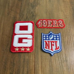 May include: Three embroidered patches featuring sports themes. One rectangular red patch displays the white letters OG with three gold stars below. A red banner patch reads 49ERS in gold and white. A blue shield patch features the NFL logo and a football.