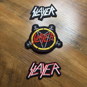 May include: Three Slayer band patches. The top patch is a black and white logo in a jagged shape. The middle patch is a black circle with a yellow border, a red Slayer logo, and a pentagram. The bottom patch is a red and white Slayer logo.