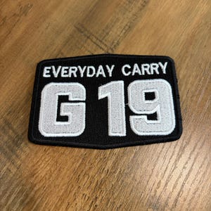 May include: A rectangular black embroidered patch with a stitched border. The text "EVERYDAY CARRY" is embroidered in white at the top, and the large white characters "G 19" are centered below, featuring a thick white outline and black fill.