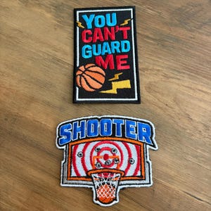 May include: Two embroidered patches on a wooden surface. The top patch reads "YOU CAN'T GUARD ME" with a basketball graphic. The bottom patch says "SHOOTER" above a basketball hoop and target design. The patches feature bright colors.
