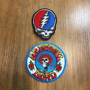 May include: Two embroidered patches. The top patch is a skull shape with a red and blue background and a white lightning bolt. The bottom circular patch features a central skull surrounded by red roses, with the text Grateful Dead in yellow.