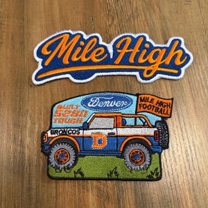 May include: Two embroidered patches. The top patch features the text Mile High in orange script on a blue background with a white border. The bottom patch depicts a blue and white off-road vehicle with orange accents, the text Denver, 5280 Tough, Broncos, and Mile High Football on a flag.