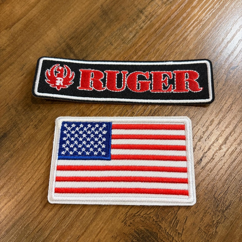 May include: Two embroidered patches. The top patch is black with the word "RUGER" in red and a red logo. The bottom patch is a rectangular American flag with red and white stripes and a blue field of white stars.