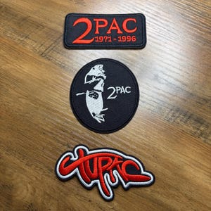 May include: Three black and red embroidered patches featuring the name "2PAC". One rectangular patch has the text "2PAC 1971-1996". A circular patch displays a portrait of a person with the text "2PAC". The third patch has the name "2PAC" in a stylized font.