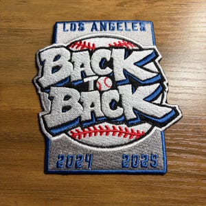 May include: Embroidered patch with the text "LOS ANGELES" at the top. The words "BACK TO BACK" are in large, stylized letters over a baseball graphic. The years "2024 2025" are at the bottom. The patch has blue, white, red, and gray colors.