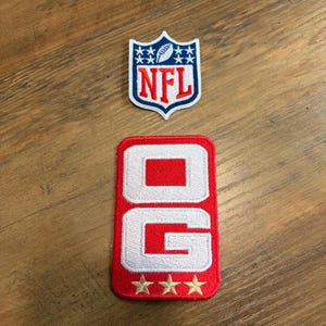May include: A shield-shaped NFL patch featuring a white football icon, blue stars, and red lettering on a white background, positioned above a rectangular red patch with white block letters reading OG and three gold embroidered stars at the base.