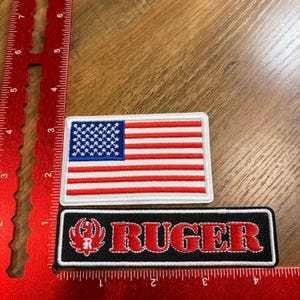 May include: Two embroidered patches. The top patch is a rectangular American flag with red and white stripes and a blue field of stars. The bottom patch is black with the word "RUGER" in red and a red logo.