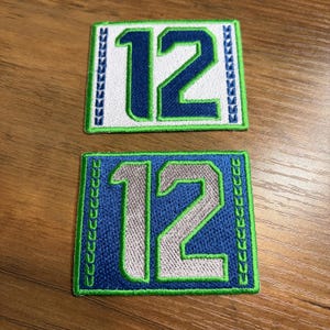 May include: Two embroidered patches with the number "12". One patch has a white background with blue and green accents. The other patch has a blue background with silver and green accents. Both patches are rectangular.