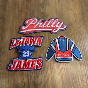May include: Three embroidered patches featuring red, white, and blue designs. One patch reads Philly in script. Another patch features a blue Liberty Bell with the text LeJawn 23 James. The third patch depicts a blue and red pullover jacket.