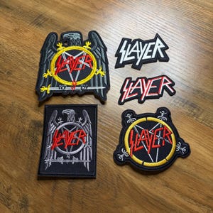 May include: Four embroidered patches with the word "SLAYER" in various designs. One patch features an eagle with a pentagram and the word "SLAYER" in red. The other patches have the word "SLAYER" in white and red.