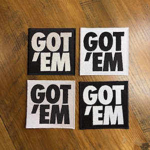 May include: Four square patches with the text "GOT 'EM" in a bold, blocky font. Two patches have a black background with white text, and two have a white background with black text. The patches are arranged in a square.