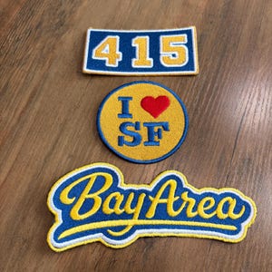 May include: Three embroidered patches featuring blue and yellow color schemes. The top rectangular patch displays the number 415. The middle circular patch shows the text I heart SF with a red heart. The bottom patch features the text Bay Area.