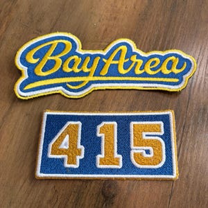May include: Two embroidered patches with blue and yellow thread. The top patch features the text Bay Area in a cursive script with a yellow underline. The bottom rectangular patch displays the number 415 in a bold, block-style yellow font.