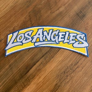 May include: Embroidered patch with the text "LOS ANGELES" in white, outlined in light blue, on a yellow banner. The patch has a blue border and is likely for a jacket or bag.