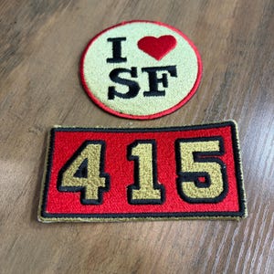 May include: A circular embroidered patch with a cream background, a red border, and the text I heart SF in black with a red heart symbol, alongside a rectangular red embroidered patch with a black border and the gold numbers 415 in black outline.