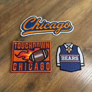 May include: Three embroidered patches featuring Chicago sports themes. One patch displays the word Chicago in blue script with orange outlines. Another is a rectangular patch with the text Touchdown Chicago and a flaming football. The third is a jersey patch.