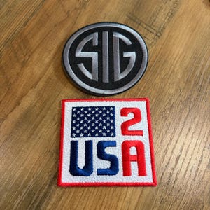May include: A circular black patch with a silver SIG logo and a square white patch featuring a blue field of stars, the blue text USA, and the red number 2. Both embroidered patches have stitched borders and are displayed on a wooden surface.