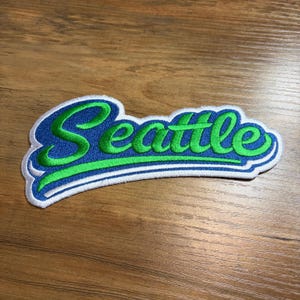 May include: Embroidered patch featuring the word "Seattle" in green script lettering, outlined in white, on a blue background. The patch has a white border and is likely for clothing or accessories.