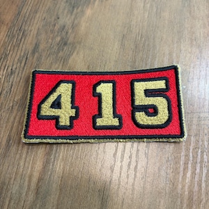 May include: A rectangular embroidered patch featuring the number 415 in gold thread with a thick black outline. The background of the patch is a vibrant red, and the entire design is bordered by a thin black stitched edge on all four sides.