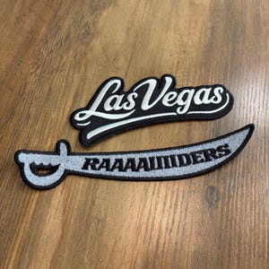May include: Two embroidered patches. The top patch features the text Las Vegas in white cursive script on a black background. The bottom patch is shaped like a curved sword with a silver blade and black handle, featuring the text RAAAAIIDERS in black.
