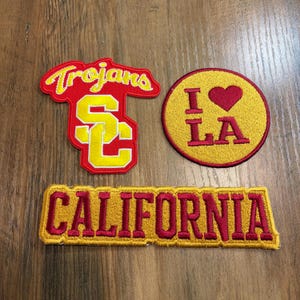 May include: Three embroidered patches: one red and yellow patch with the text Trojans and the letters SC, one circular yellow patch with a red heart and the text I Love LA, and one rectangular yellow patch with the red text CALIFORNIA.