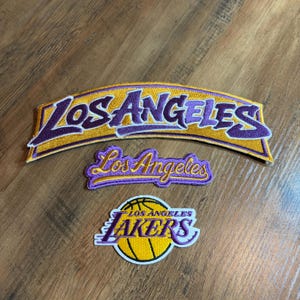 May include: Three embroidered fabric patches featuring Los Angeles Lakers branding. The top patch is a large, arched gold and purple Los Angeles text. The middle patch is a smaller, cursive Los Angeles script. The bottom patch is a circular basketball logo.