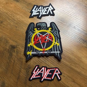 May include: Three Slayer band patches. The top patch features the band's name in white, outlined in black. The middle patch is a black eagle with a yellow and red Slayer logo. The bottom patch has the Slayer name in red and white.