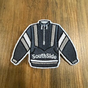 May include: An embroidered patch shaped like a dark gray pullover jacket with a white zipper, white stripes on the sleeves, and the word SouthSide stitched in white lettering across the front. The patch features a white border and detailed stitching.
